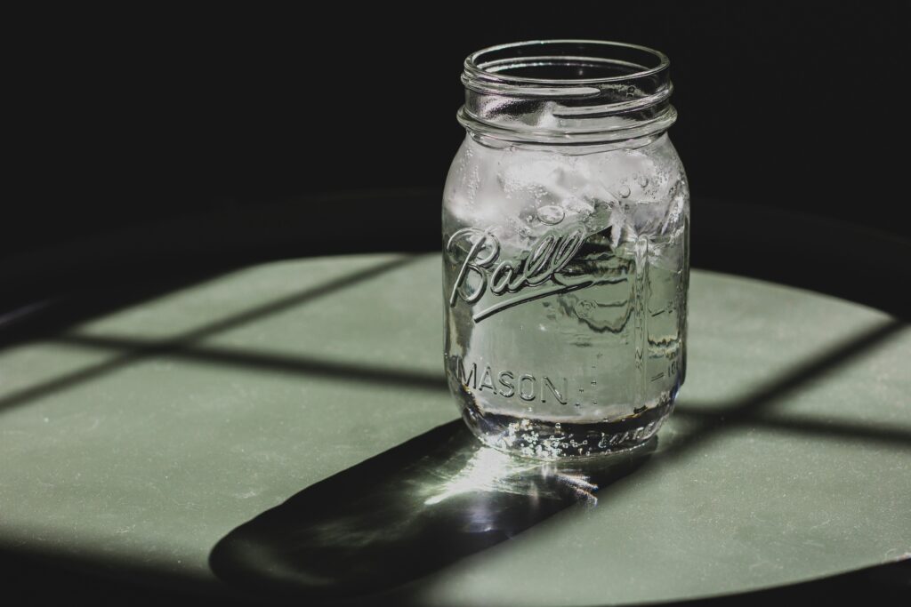 Close-up of a clear Ball mason jar filled with ice water, casting a shadow on a green table, captured in natural light.