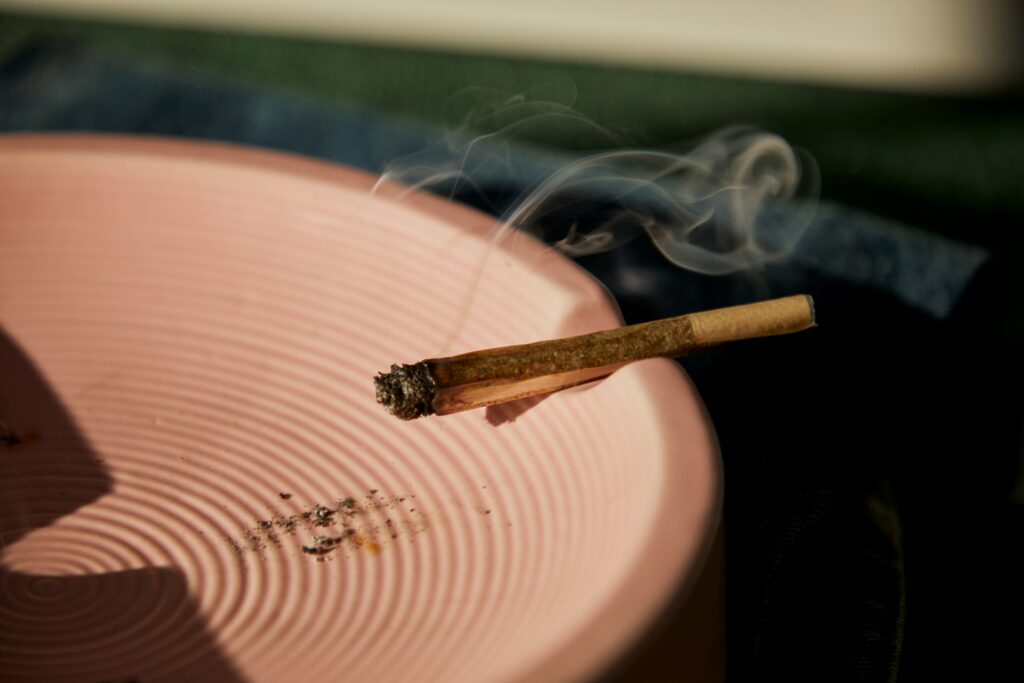 A close-up image of a lit cannabis joint resting on the edge of a pink ceramic ashtray. The joint's tip is slightly burned, with a trail of smoke rising gently into the air. The ashtray has a concentric circular pattern, adding texture to the scene. The overall lighting gives the image a warm, relaxed atmosphere, suggesting a moment of calm or reflection.