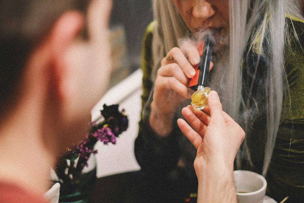 A close-up shot of two people engaging in a cannabis consumption session. One person is holding a small jar of yellow cannabis concentrate, while the other person uses a vaporizer device to inhale the vapor produced from the concentrate. The scene is set in an intimate, casual environment with some out-of-focus elements in the background, such as a plant with purple flowers. The image captures a moment of shared experience and relaxation.
