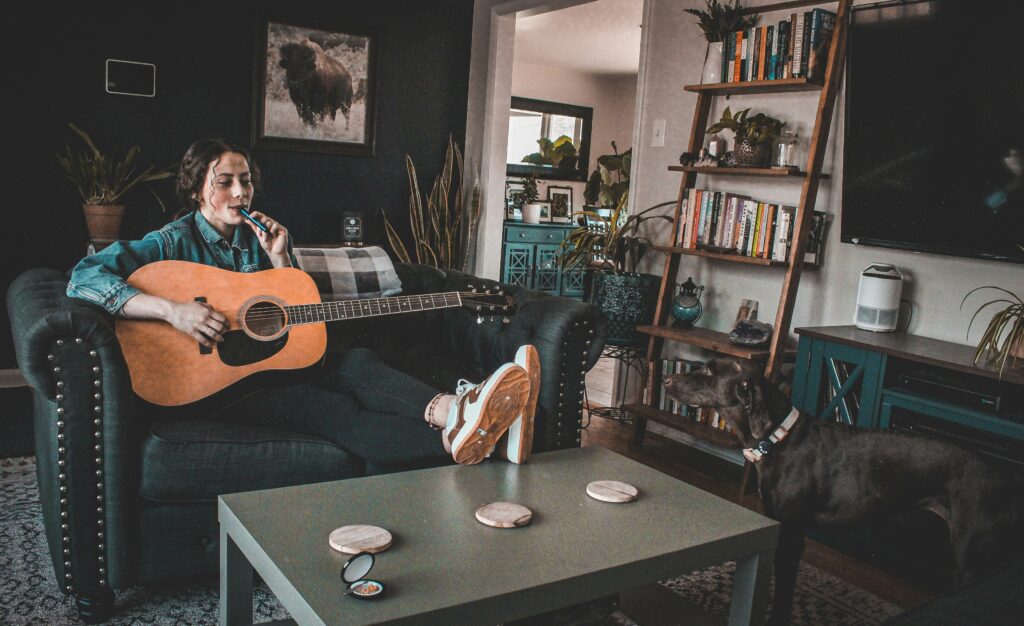 A woman lounges on a couch, playing an acoustic guitar while vaping, with a dog standing nearby in a cozy, book-filled living room. This image captures a relaxed, homey atmosphere with elements of music, relaxation, and companionship.