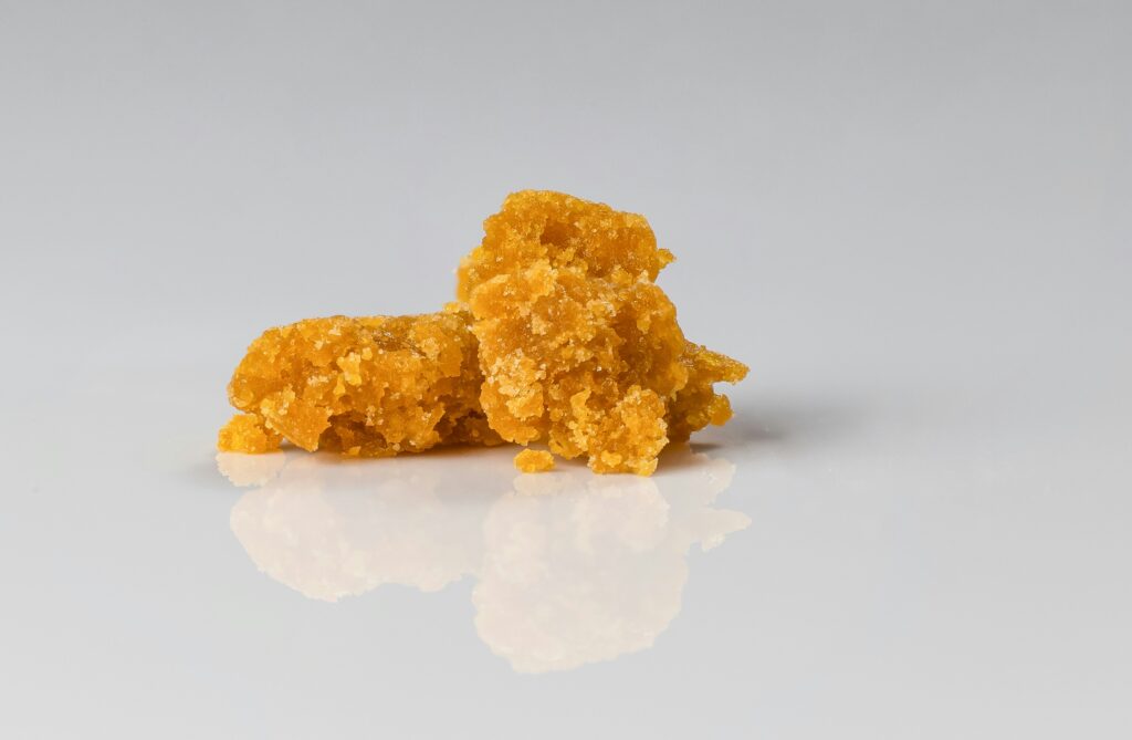 A close-up image of a piece of cannabis concentrate, likely a crumble or wax, displayed on a reflective white surface. The concentrate is a rich golden-yellow color, with a crumbly texture that shows its drier consistency. The reflection on the surface beneath adds depth to the image, highlighting the product's quality and texture.