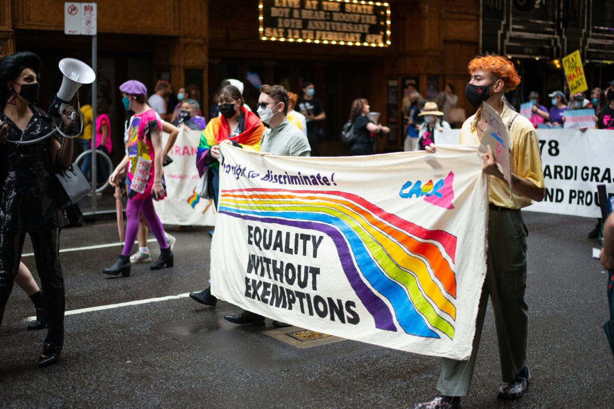A vibrant LGBTQ+ pride march featuring participants holding a banner with a rainbow design that reads 'Equality Without Exemptions.' The scene captures the energy and diversity of the march, with individuals wearing masks, colorful clothing, and carrying signs advocating for equal rights and non-discrimination.