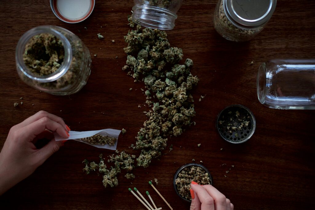 A person's hands expertly rolling a cannabis joint, surrounded by scattered cannabis buds and jars on a wooden table, illustrating the personal ritual of preparing cannabis for consumption.