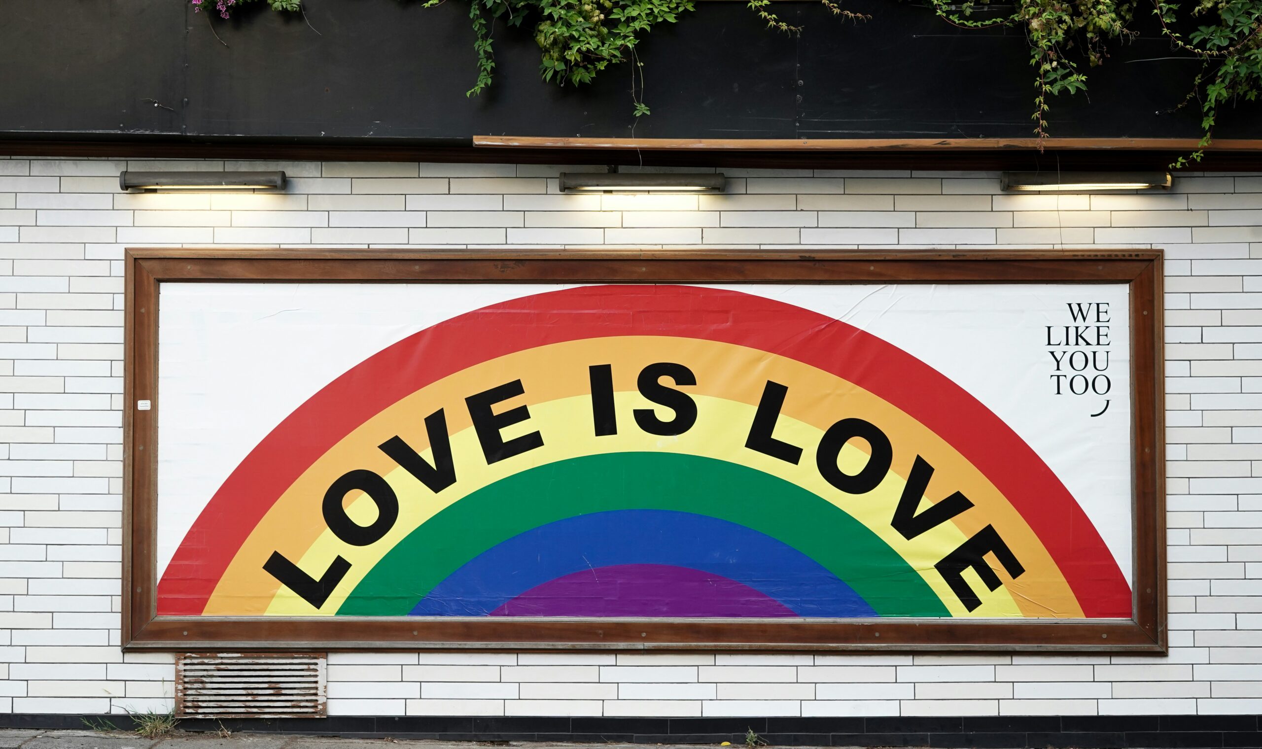 A mural featuring a vibrant rainbow arch with the bold text 'LOVE IS LOVE' prominently displayed across it, symbolizing LGBTQ+ pride and equality. The mural is framed against a white-tiled wall, with a small message 'WE LIKE YOU TOO' written on the side.