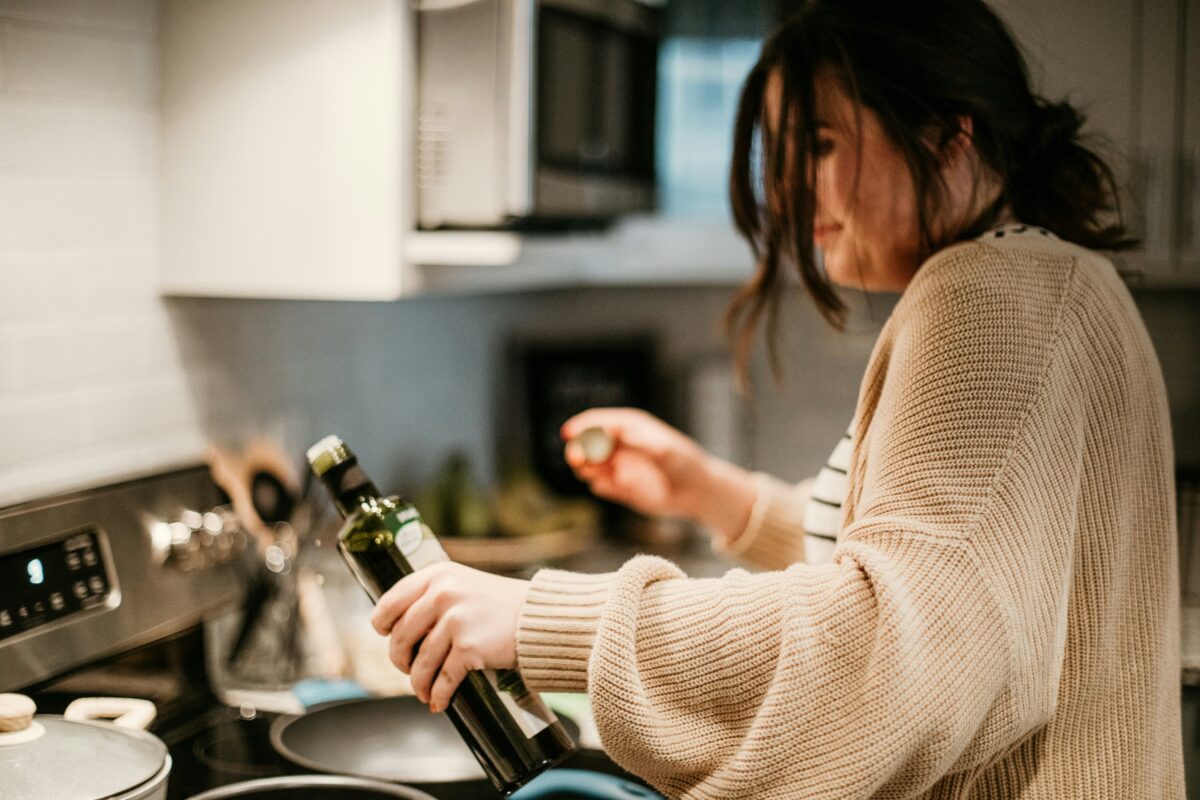 A woman with dark hair pulled into a loose bun is cooking in a kitchen. She is holding a bottle of olive oil in one hand and a bottle cap in the other, as she prepares to pour the oil into a pan on the stove. She is wearing a beige knit cardigan over a striped shirt, with a stovetop and various kitchen utensils visible in the background.