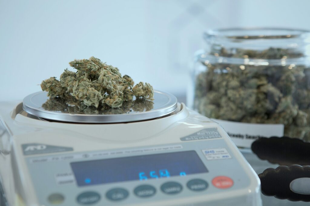 A close-up view of cannabis buds being weighed on a digital scale. The scale's display shows a measurement, while more cannabis buds are visible in a glass jar in the blurred background. The scene takes place in a well-lit, clean environment, likely in a dispensary or a lab setting.