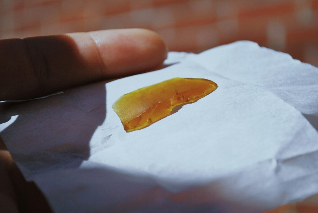 A close-up shot of a hand holding a piece of amber-colored cannabis concentrate, placed on a piece of white parchment paper. The concentrate has a smooth, glassy texture and is partially translucent, with light shining through it. The background is blurred, focusing attention on the concentrate and the hand holding it.