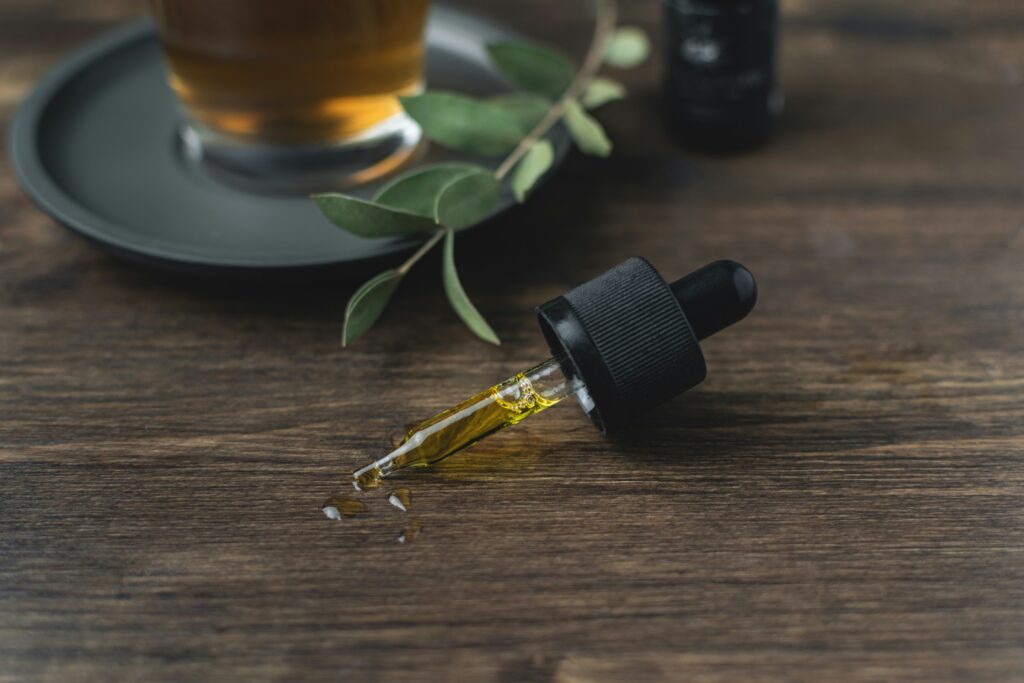 A close-up of a dropper filled with golden oil, resting on a wooden surface. A few drops of the oil are spilled beside the dropper. In the background, there is a glass cup with a dark beverage placed on a saucer, along with a small sprig of greenery. The scene has a warm, natural feel, suggesting the use of the oil for wellness or relaxation purposes.