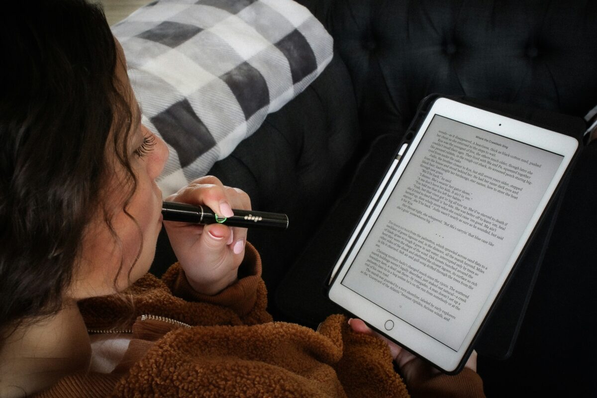 A close-up of a woman relaxing on a dark-colored couch while using a vape pen. She is holding an iPad or tablet in one hand, reading text displayed on the screen. The woman is wearing a brown fleece jacket, and a black and white checkered blanket is draped over the couch beside her. The setting appears cozy and comfortable, emphasizing relaxation and leisure.