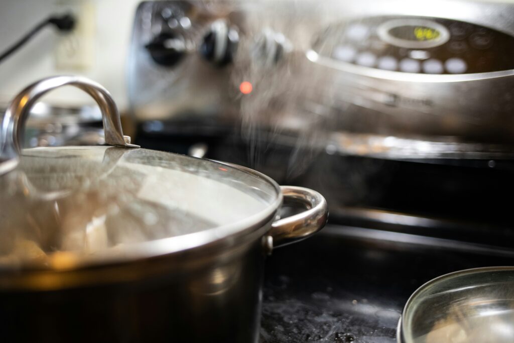 A close-up view of a stainless steel pot with a glass lid, placed on a stove. Steam is visibly rising from the pot, indicating that something is being cooked inside. The background shows a blurred view of a modern stovetop with control knobs and a digital display.