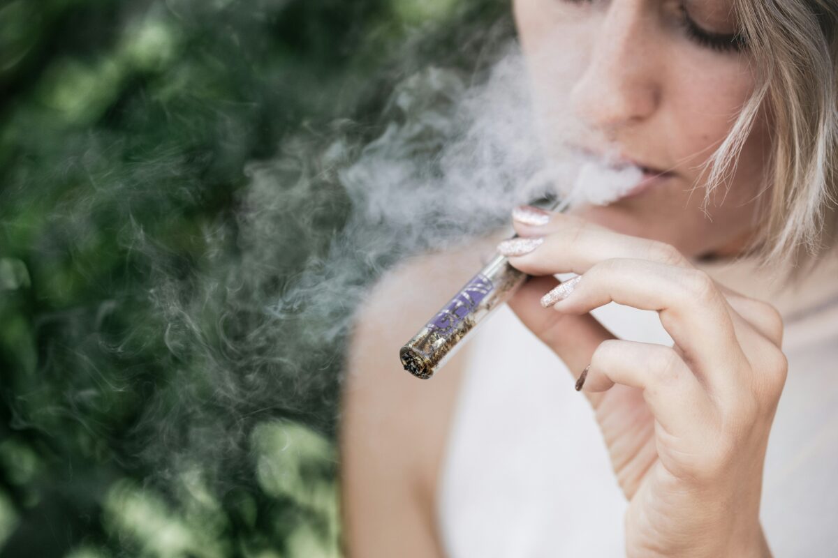 A close-up of a person inhaling from a glass smoking pipe, with visible smoke swirling around them. The person has light-colored hair and glittery nail polish, and the background is blurred with greenery, creating an outdoor setting. The image captures the act of smoking, emphasizing the calm and reflective mood.