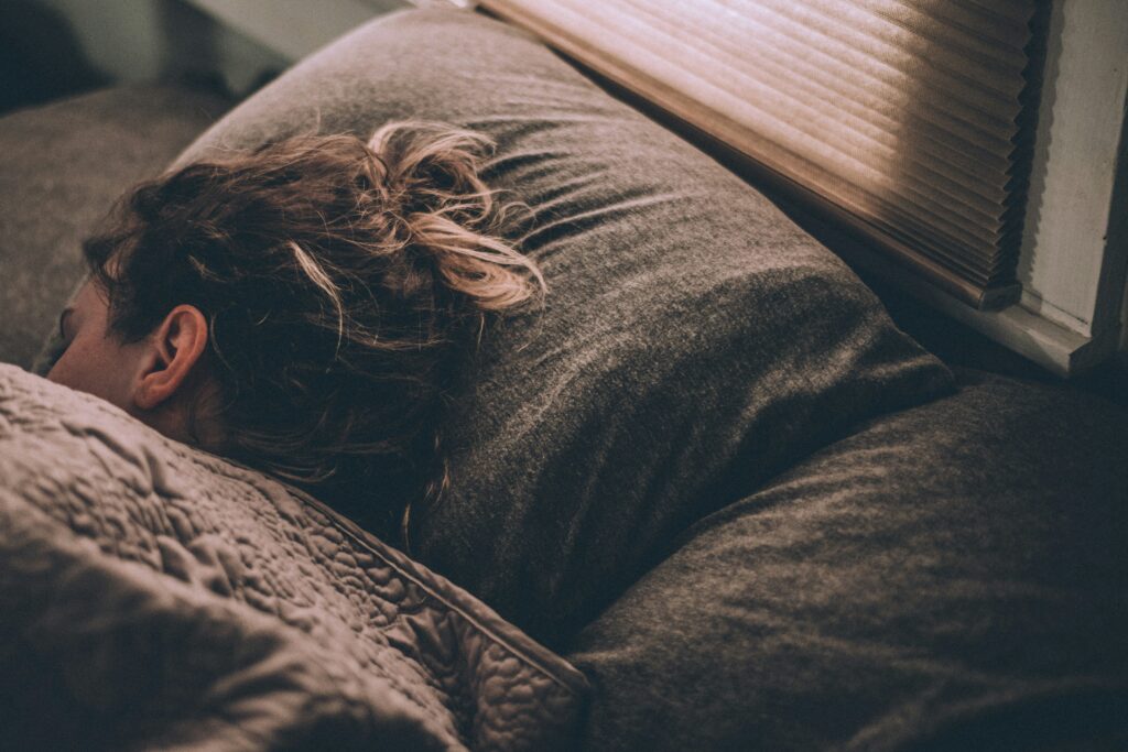 A close-up shot of a person sleeping in a bed, with their head resting on a gray pillow. The person's hair is tied up loosely, and they are covered with a soft, textured blanket. The scene is calm and serene, with soft lighting filtering through window blinds in the background, creating a peaceful atmosphere.