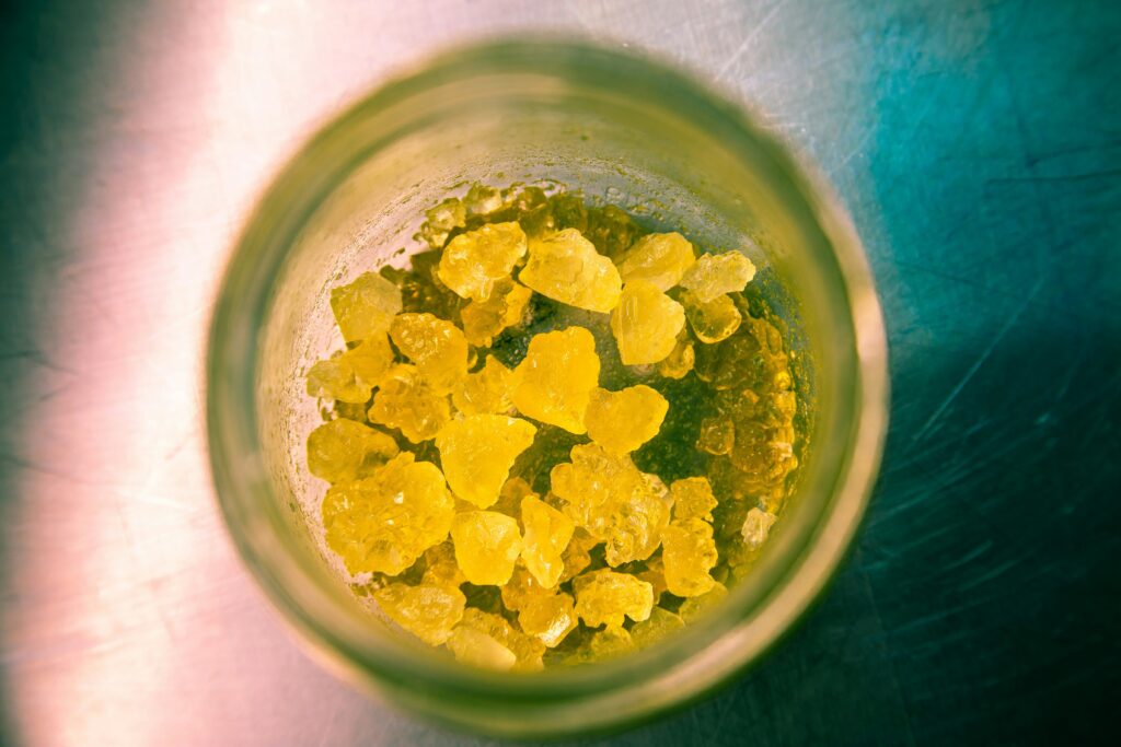 A top-down view of a glass jar containing bright yellow cannabis concentrates or crystals. The vibrant yellow crystals are clustered together, and the jar is illuminated by natural light, highlighting the texture and color of the concentrates. The background is a blurred, metallic surface, adding contrast to the vivid yellow contents of the jar.