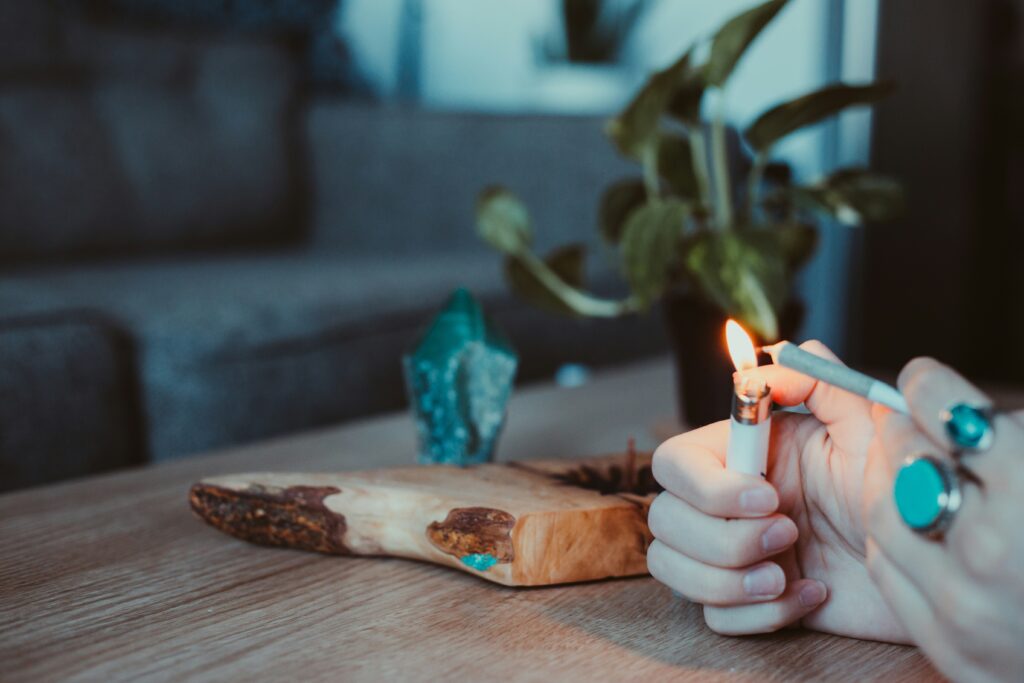 A close-up of a hand holding a lit lighter, igniting the end of a pre-rolled joint. The hand is adorned with turquoise rings. The scene is set in a cozy indoor environment with a blurred background that includes a wooden table, a decorative crystal, and a potted plant, giving the setting a relaxed and intimate vibe.