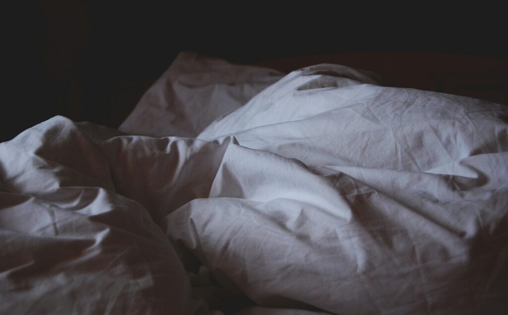A close-up shot of a rumpled white bedspread in a dimly lit room. The soft light casts gentle shadows on the fabric, creating a peaceful and serene atmosphere. The scene evokes a sense of comfort and restfulness, with the inviting, unmade bed suggesting a space for relaxation or sleep.