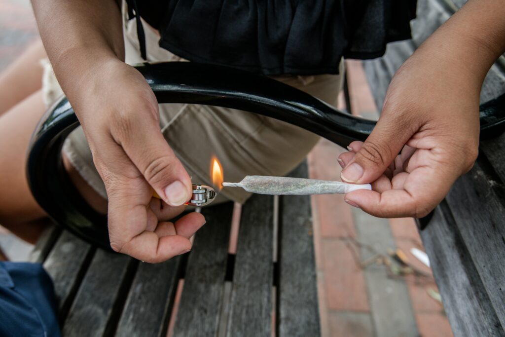 A close-up shot of a person sitting on a bench, lighting a joint with a small lighter. The person is holding the joint with one hand while using the other to ignite it. The individual is dressed in casual clothing, including light-colored shorts and a black top. The scene takes place outdoors, with a park or public space setting visible in the background.