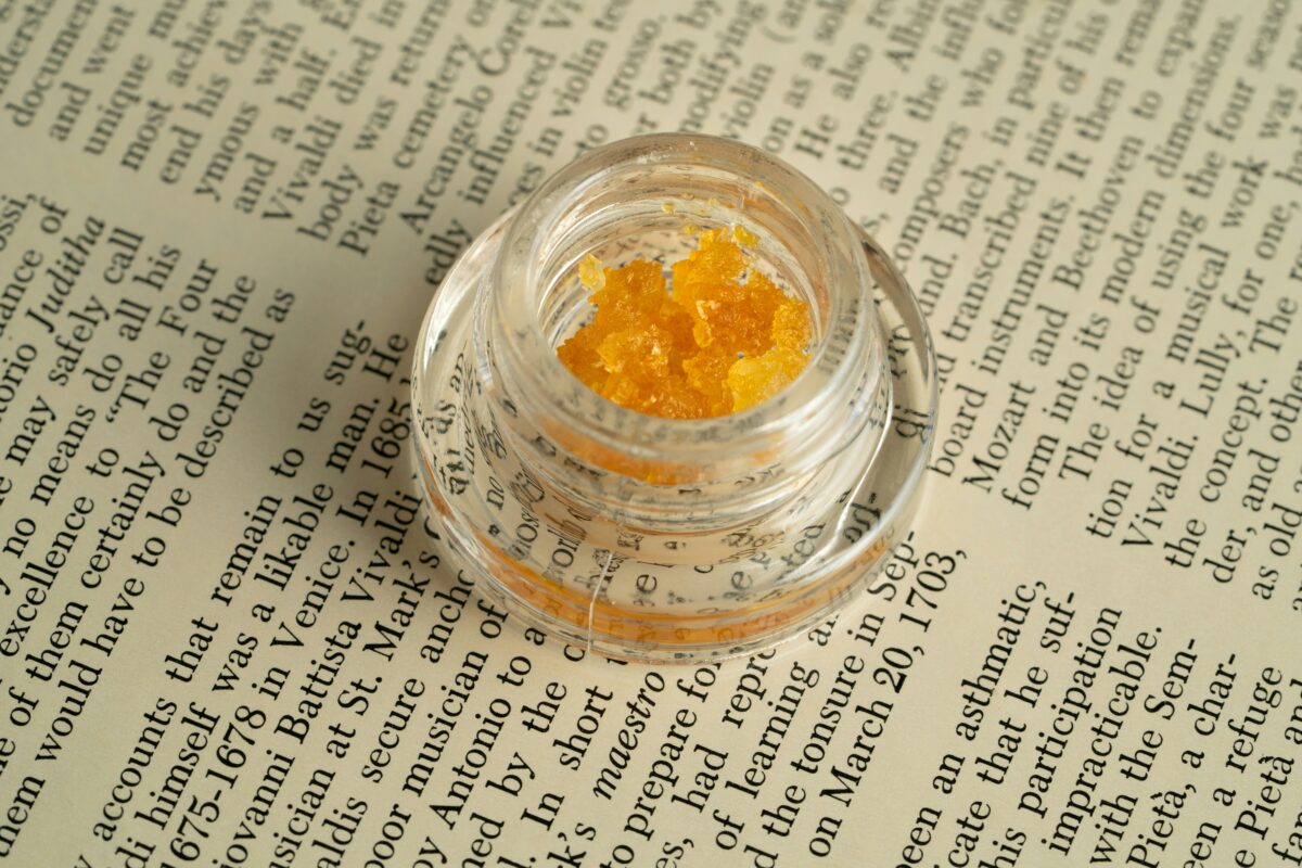 A small, clear jar containing bright yellow cannabis concentrates or crystals is placed on top of an open book with text visible on the pages. The jar is slightly open, revealing the textured concentrates inside. The juxtaposition of the cannabis product with the printed text creates an interesting contrast between modern cannabis use and traditional printed material.