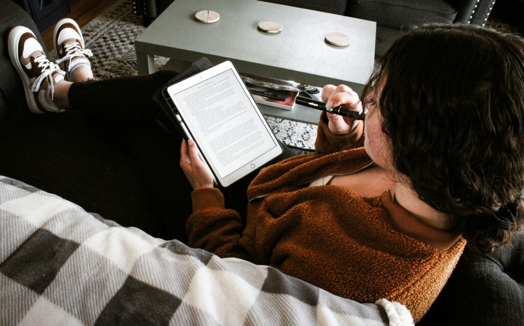 A person with dark, wavy hair sitting on a couch, holding a tablet with both hands, reading text displayed on the screen. They are wearing a brown fleece jacket and black leggings, with their legs crossed. The person is using a vape pen, with the vape close to their mouth. A coffee table with coasters and other items is in the background.