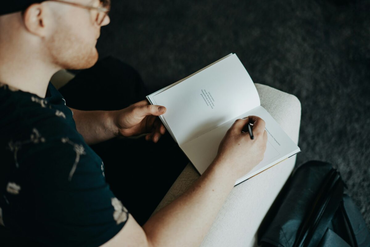 A person with a short beard and glasses writing in an open notebook, sitting on a light-colored chair. The notebook has a small amount of printed text at the top of the page, with the person holding a black pen ready to write. The background is out of focus, highlighting the writing action.