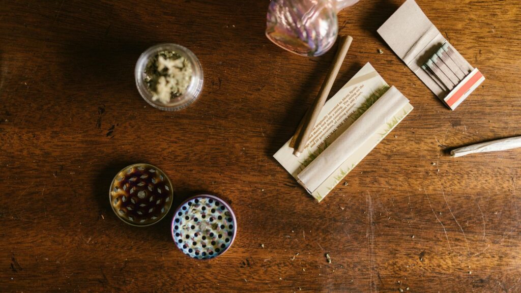 5 - Nature Med Dispensaries Overhead view of a wooden table with cannabis preparation items, including a glass jar with flower, colorful grinder, rolling papers, matches, and a rolled pre-roll, capturing a relaxed setup for rolling.