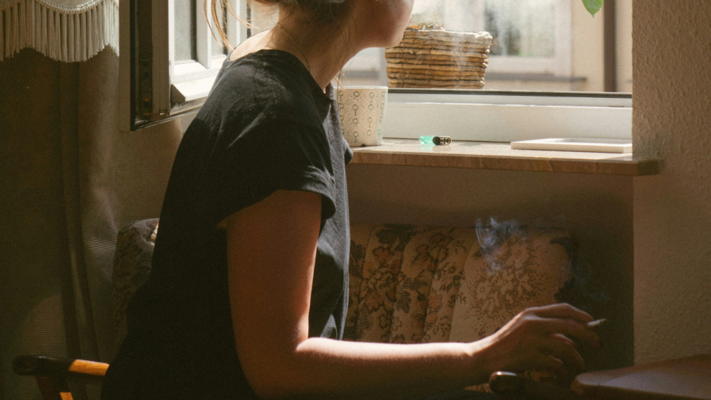 Person relaxing by an open window, holding a lit pre-roll with smoke rising gently in a cozy indoor setting. Sunlight filters through, illuminating a cup and lighter on the windowsill, creating a peaceful, reflective atmosphere.