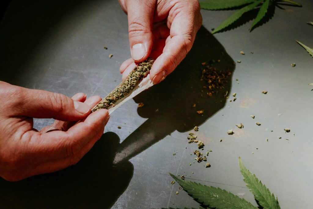 Hands carefully rolling a cannabis pre-roll on a metal surface, with scattered herb and cannabis leaves nearby, capturing the art of rolling in a relaxed setting.
