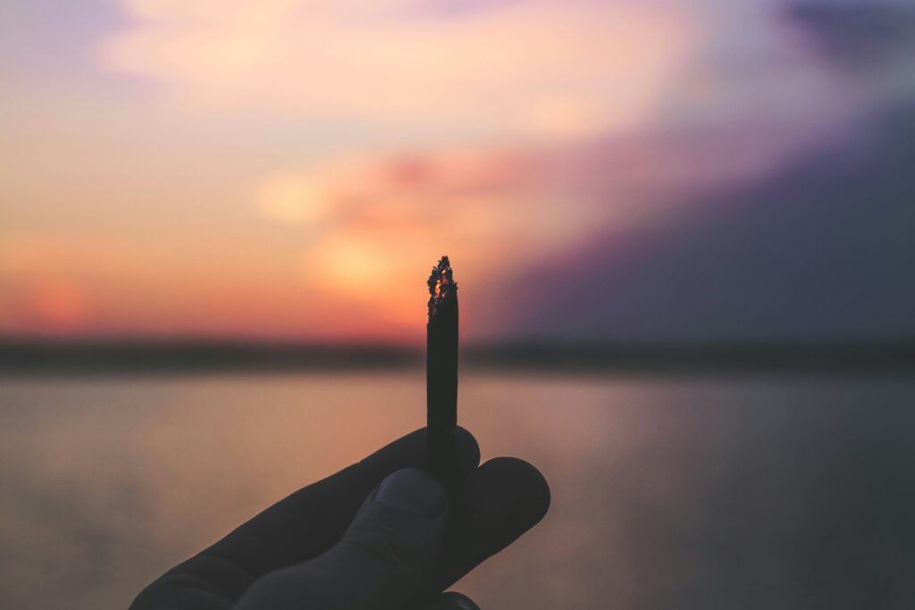 Hand holding a lit cannabis pre-roll against a serene lakeside sunset, with warm pink and purple hues in the sky creating a peaceful, reflective vibe.