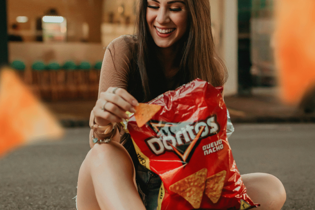 image of a woman with brown hair sitting on the ground outside with a bag of Doritos smiling.
