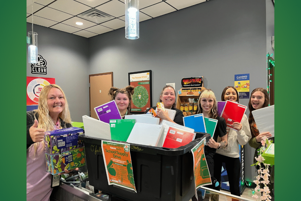 A group of five women smiling and holding a large black bin overflowing with notebooks and binders for a school supply drive. A woman on the far left holds a box of school supplies and gives a thumbs-up.