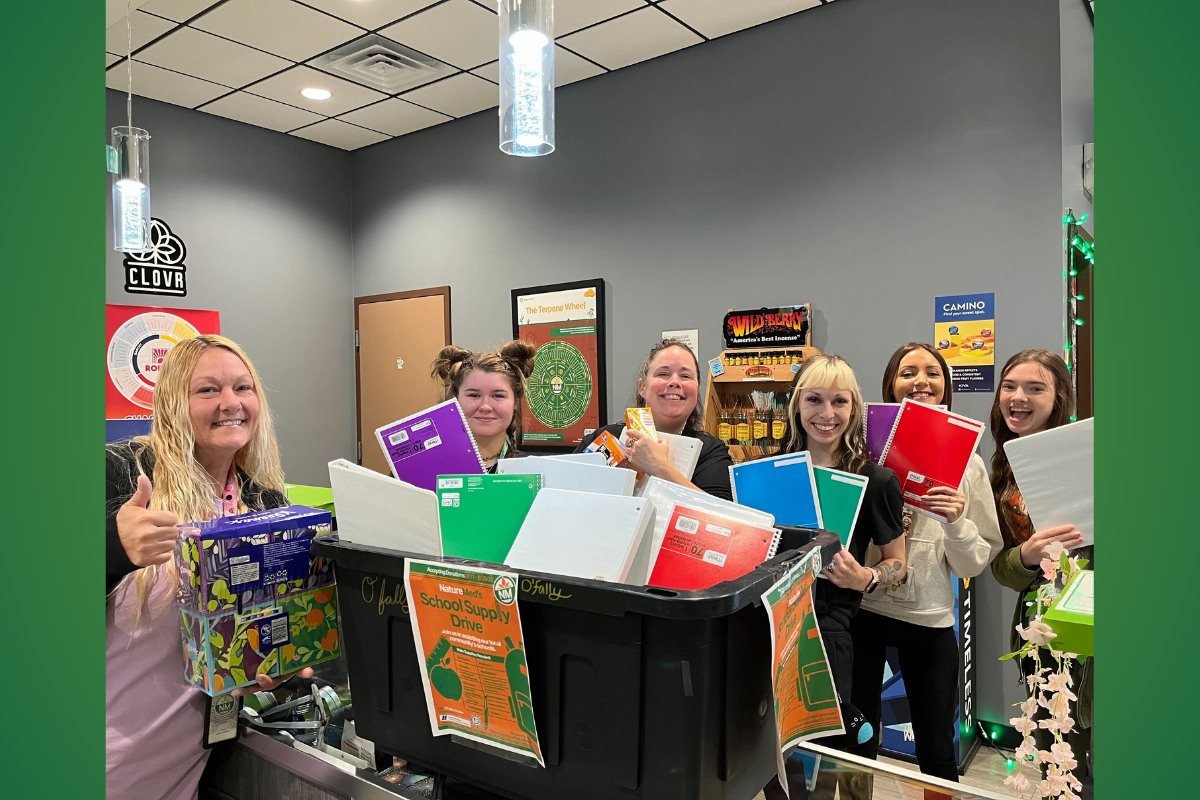 A group of five women smiling and holding a large black bin overflowing with notebooks and binders for a school supply drive. A woman on the far left holds a box of school supplies and gives a thumbs-up.