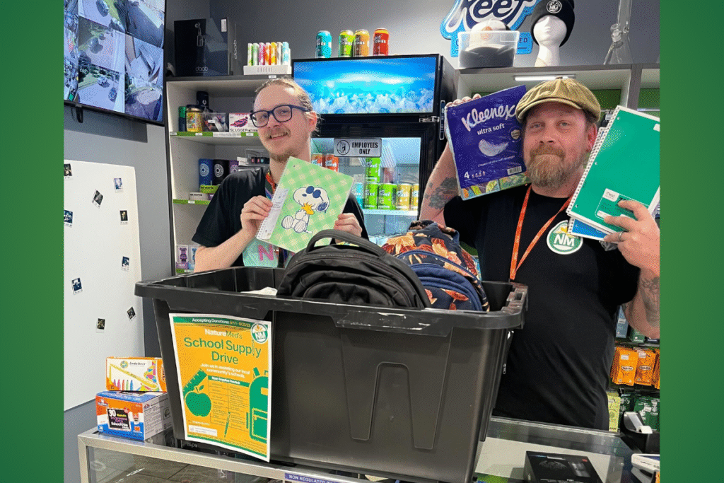 - Nature Med Dispensaries Two men pose behind a black bin filled with backpacks. One man on the left holds a notebook and the man on the right holds a box of Kleenex and notebooks. A sign on the bin says "School Supply Drive."