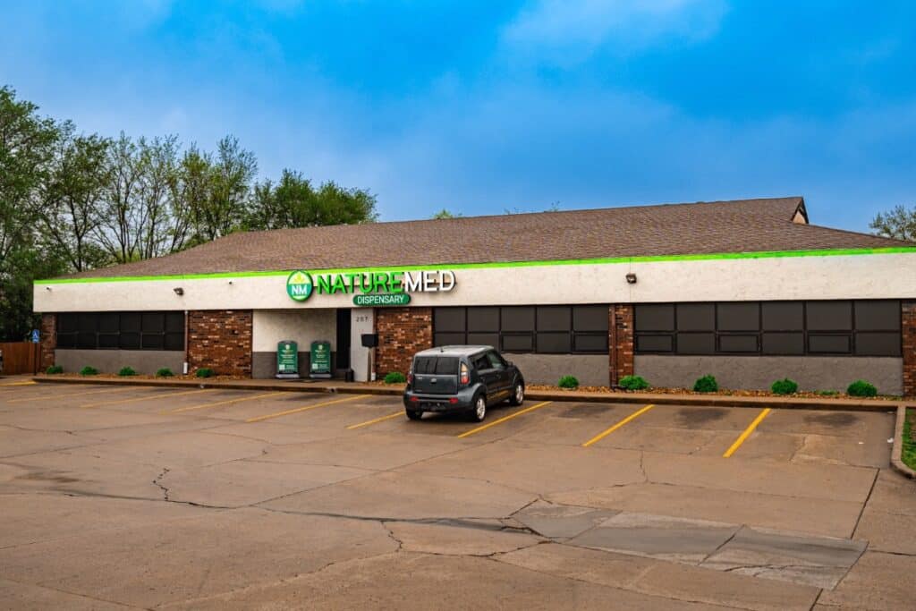 An exterior view of the NatureMed Dispensary storefront, a one-story brick building with a large sign and an empty parking lot on a sunny day.