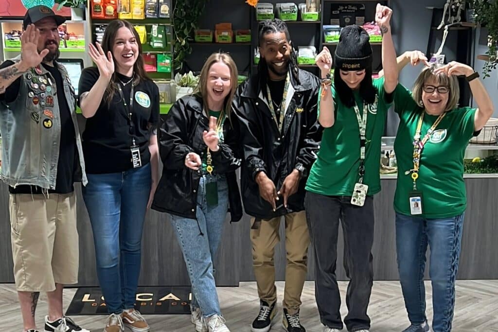 A diverse team of six happy cannabis dispensary employees wearing green branded shirts and hoodies, posing together for a group photo inside the store.