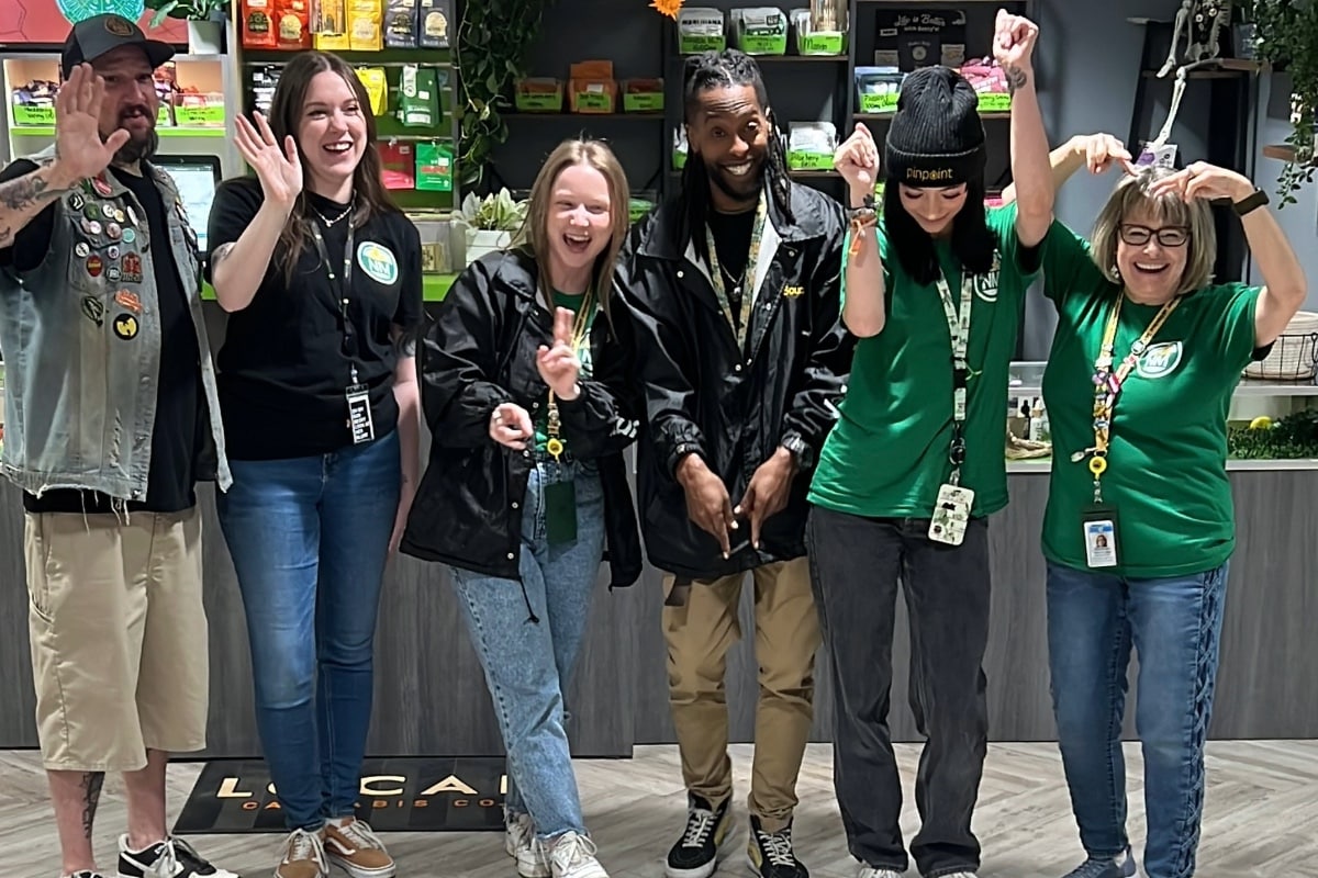 A diverse team of six happy cannabis dispensary employees wearing green branded shirts and hoodies, posing together for a group photo inside the store.