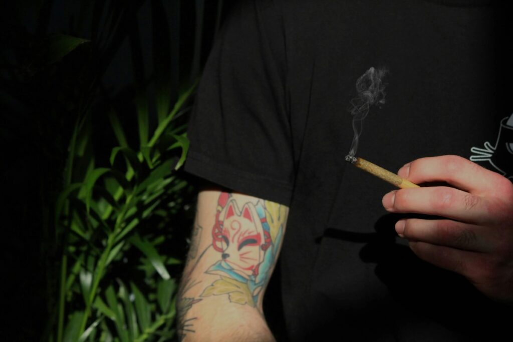 Close-up of a person's tattooed arm (featuring a kitsune mask design) holding a lit cannabis pre-roll or joint, with smoke rising from the tip. This illustrates the traditional method of smoking marijuana.