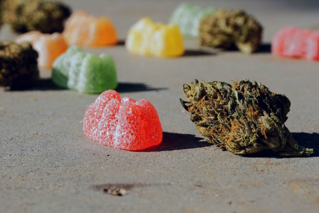 A close-up, outdoor shot featuring a red cannabis gummy edible next to a dense marijuana flower bud on a concrete surface. Out-of-focus colorful gummies and flower buds are in the background, representing the variety of cannabis edibles available.