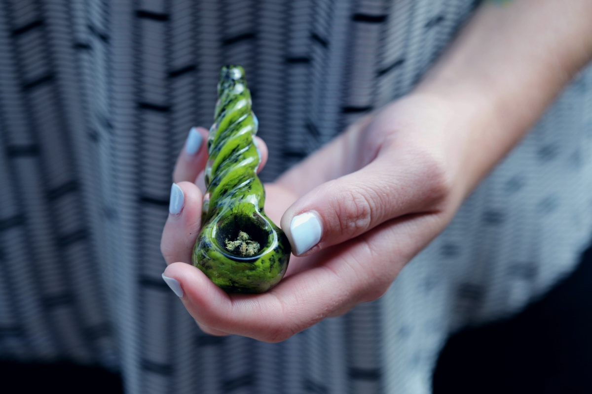 Hand holding a green glass pipe filled with ground cannabis flower. The hand has light blue nail polish, and the pipe is a unique, spiral-shaped design, highlighting smoking accessories for marijuana.