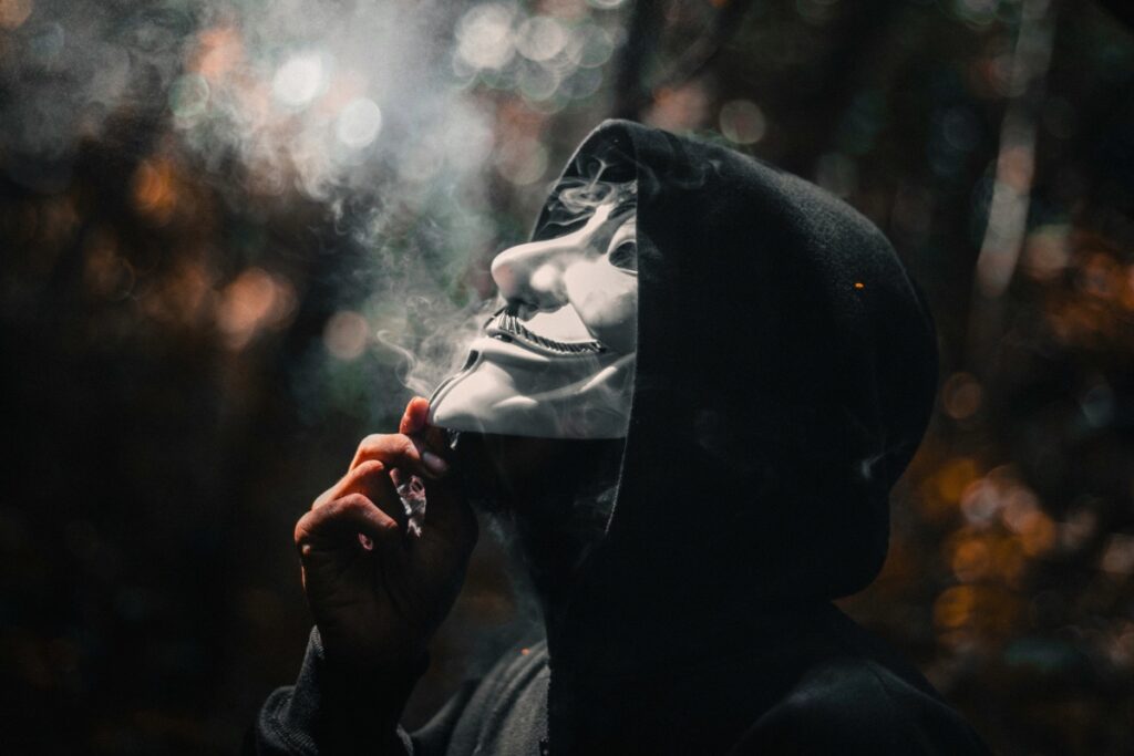 A mysterious figure in a hoodie and Guy Fawkes mask exhales a plume of smoke, perfect for a cannabis culture or hacker Halloween costume.