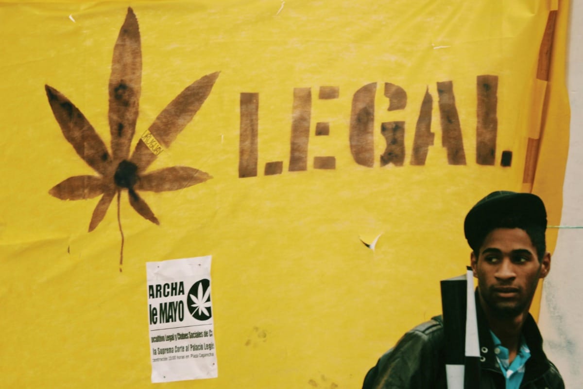 cannabis rescheduling blog-2 - Nature Med Dispensaries A man in a leather jacket stands in front of a large yellow banner featuring a brown cannabis leaf silhouette and the word "LEGAL" in bold, black block letters. A smaller flyer for a "Marcha de Mayo" event is pinned below.