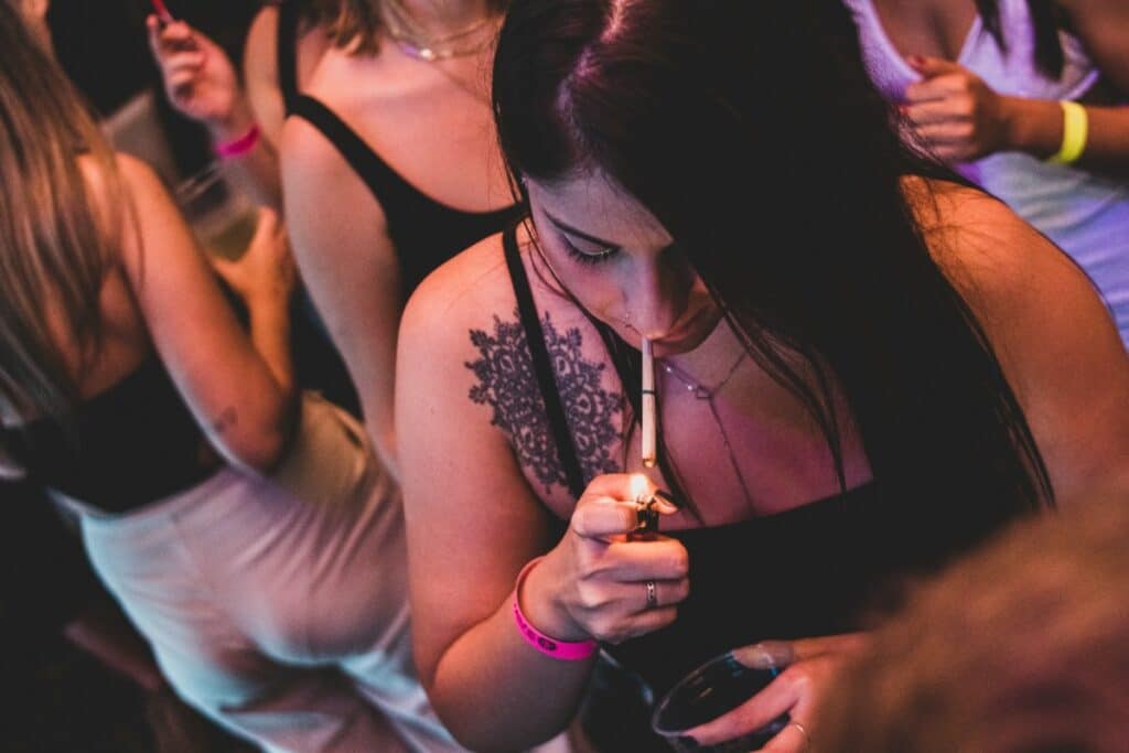 - Nature Med Dispensaries A woman lighting a cannabis pre-roll at a crowded New Year's Eve party with atmospheric pink and purple lighting.