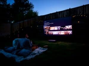 A couple enjoying a cannabis-friendly backyard movie night on a blanket with pillows, watching a large projector screen under the stars.