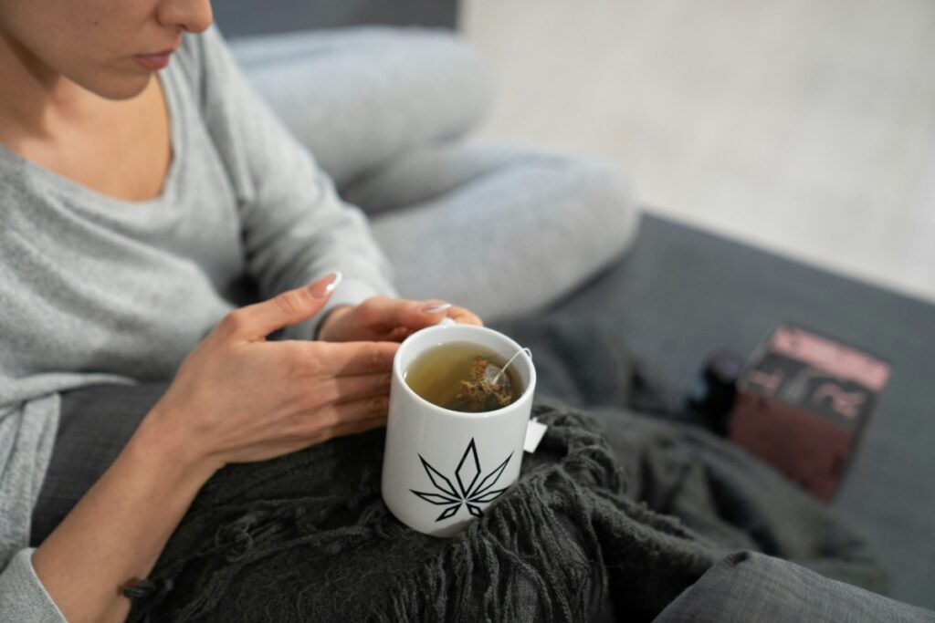 - Nature Med Dispensaries A woman relaxing on a couch wrapped in a warm blanket, holding a white mug featuring a black cannabis leaf logo while drinking herb-infused tea as part of a calming nighttime cannabis wellness routine.