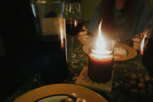 A cozy, candlelit dinner scene featuring a bottle of red wine, wine glasses, and a bowl of olives. A bright candle flame illuminates the table where a person is enjoying a meal in a dimly lit, romantic setting.