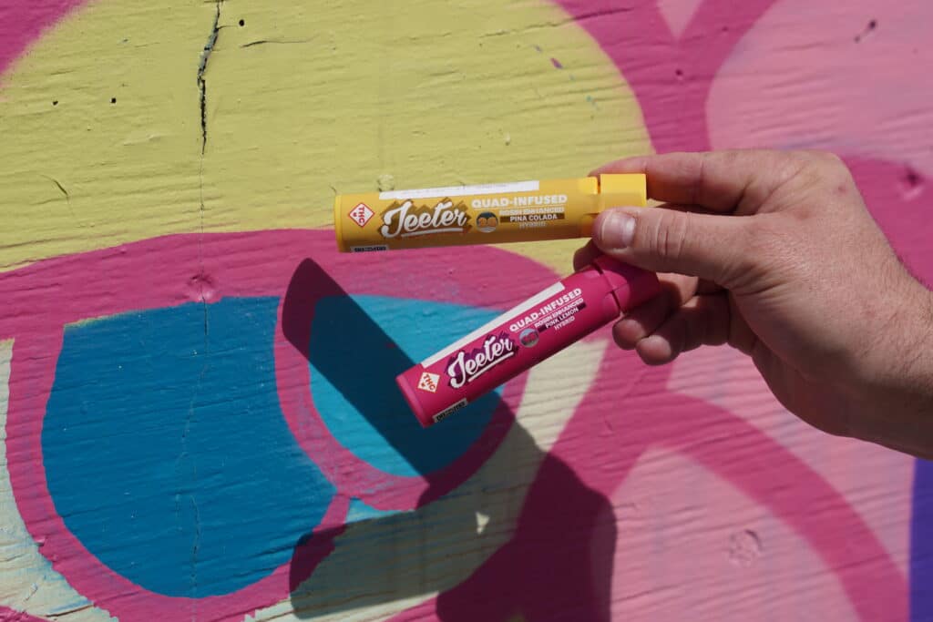 - Nature Med Dispensaries A close-up of a person’s hand holding two Jeeter tubes horizontally against a vibrant yellow, pink, and blue graffiti mural. The top tube is yellow (Piña Colada Hybrid) and the bottom is pink (Pink Lemon Hybrid).