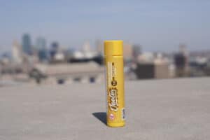 A single yellow Maui Wowie Sativa Jeeter Quad-Infused tube standing upright on a concrete ledge. In the soft-focus background, a sprawling city skyline and urban buildings are visible under a clear blue sky.