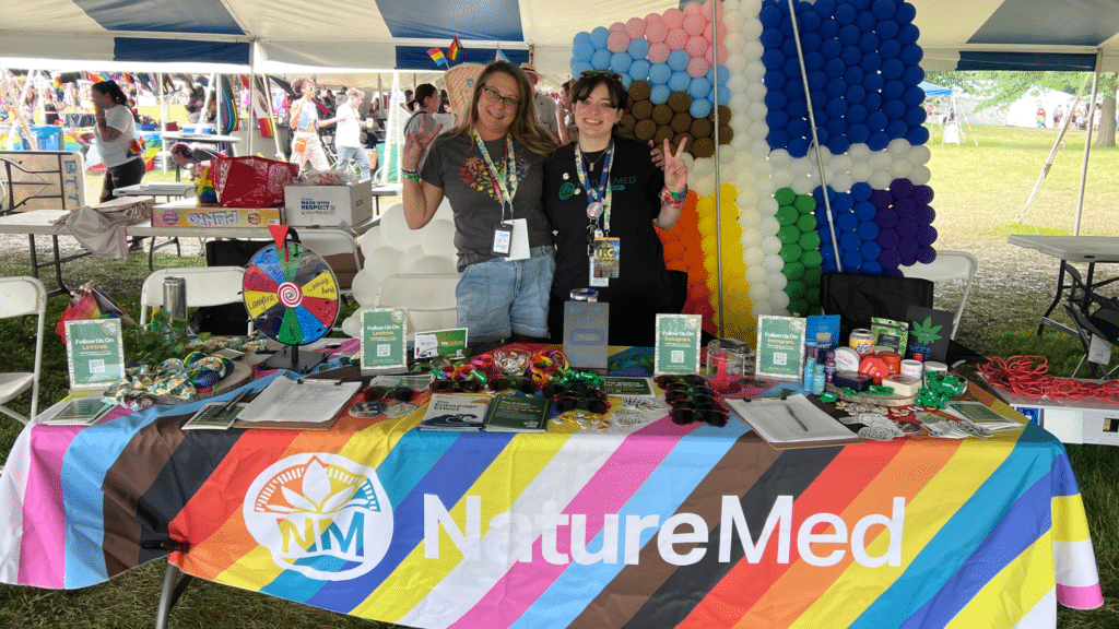 - Nature Med Dispensaries - Nature Med Dispensaries Two smiling staff members at the NatureMed cannabis dispensary vendor booth during KC PrideFest 2026, featuring a colorful branded rainbow tablecloth, a prize wheel, and festival swag.