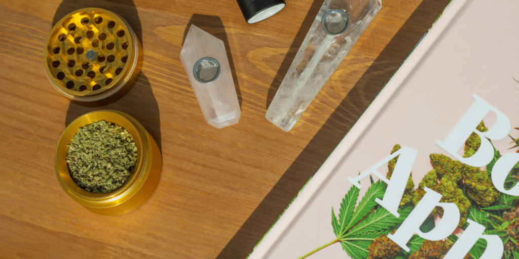 - Nature Med Dispensaries - Nature Med Dispensaries Gold herb grinder filled with ground cannabis flower sitting next to crystal pipes and a cannabis book on a wooden table.