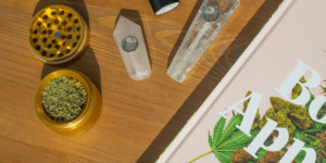 Gold herb grinder filled with ground cannabis flower sitting next to crystal pipes and a cannabis book on a wooden table.