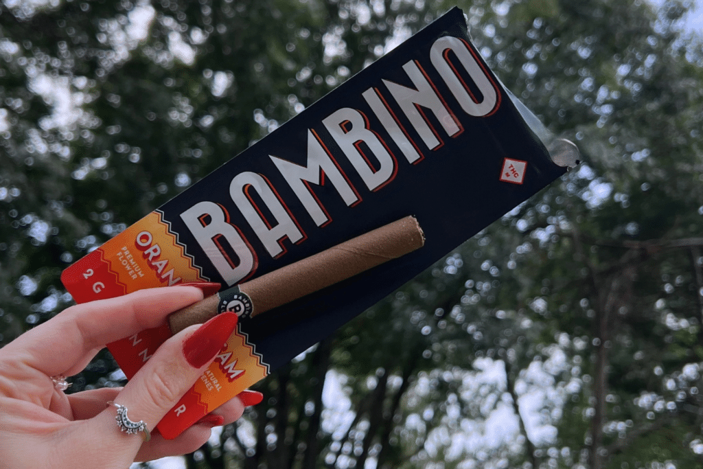 - Nature Med Dispensaries - Nature Med Dispensaries Hand holding a 2g Bambino Orange Cream pre-roll blunt over its black and orange packaging against autumn trees.