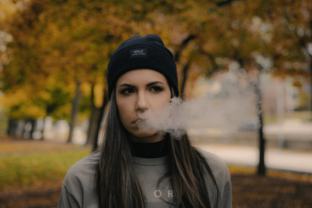 - Nature Med Dispensaries - Nature Med Dispensaries Woman in a black beanie exhaling smoke outdoors against autumn trees during a fall smoke session.